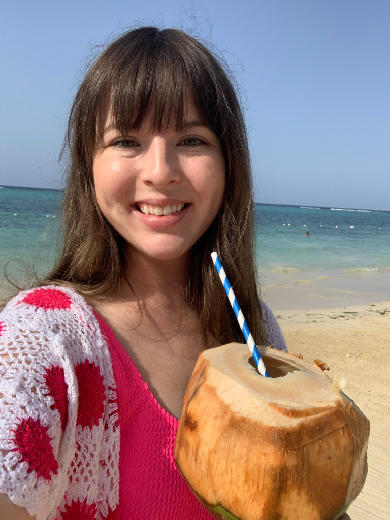 A certified travel Advisor enjoying a fresh coconut drink on a tropical beach with turquoise water in the background