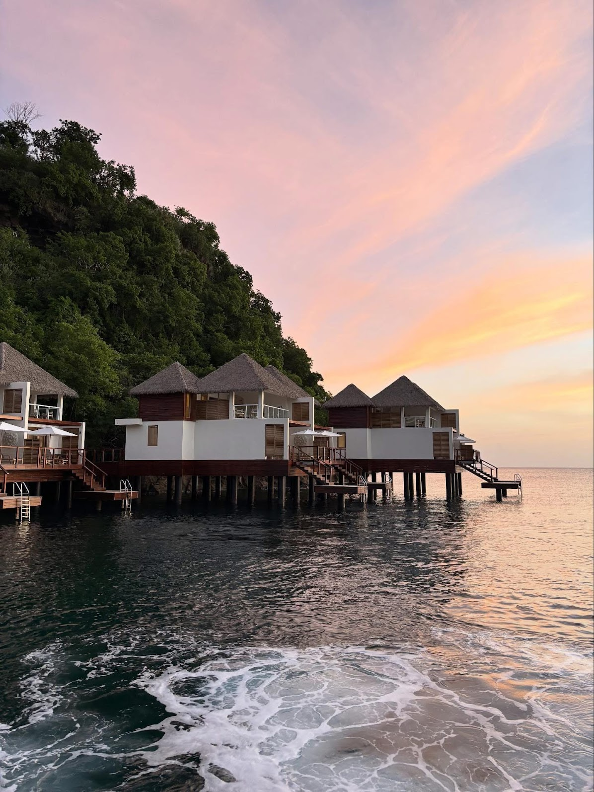 Overwater bungalows at sunset in Saint Lucia with calm ocean water and tropical hillside