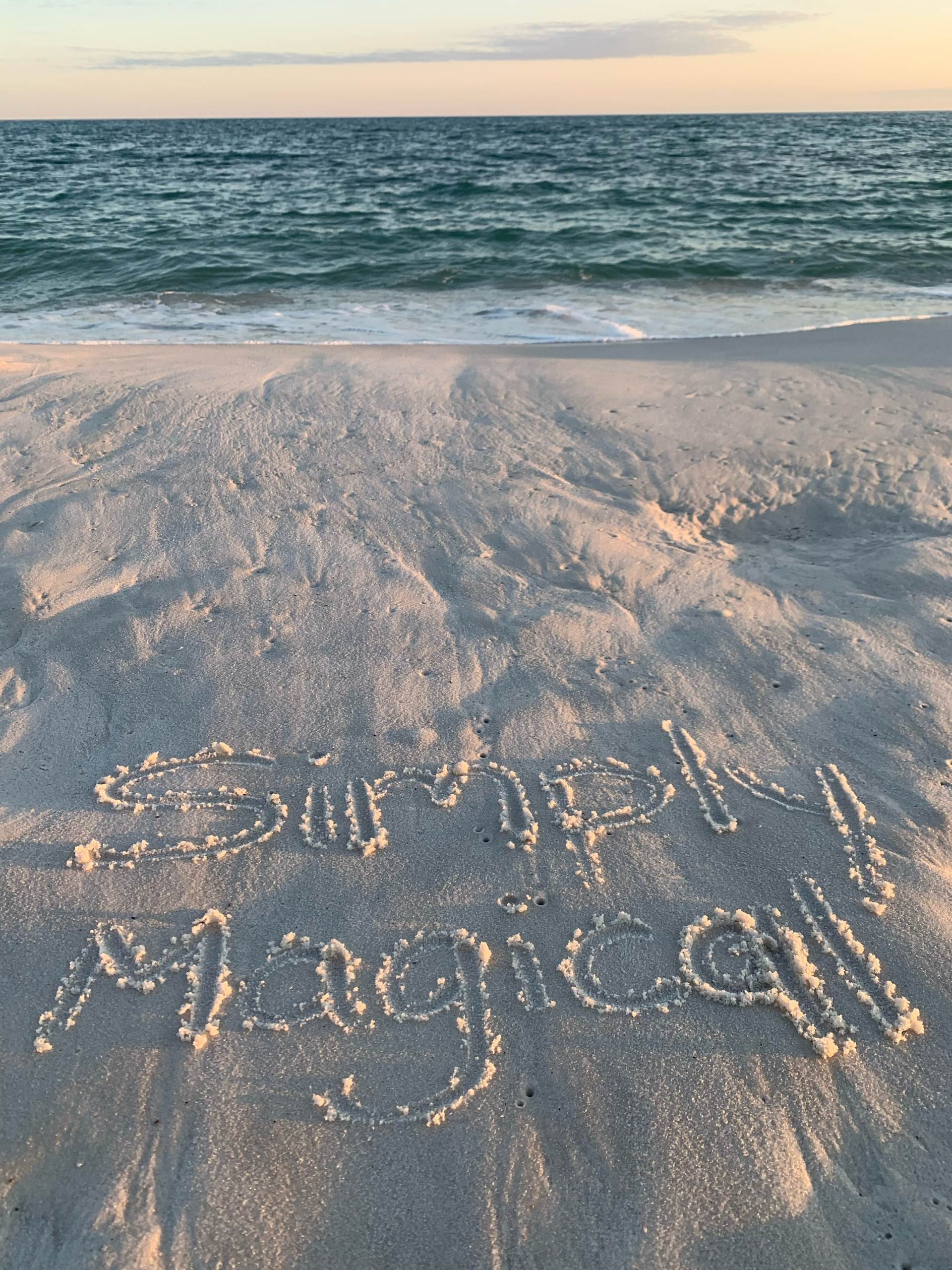 “Simply Magical” written in the sand on a beach at sunset with ocean waves in the background