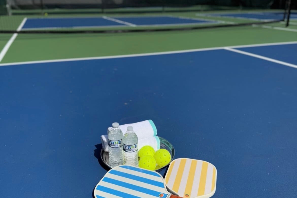 Pickleball paddles, balls, towels, and water bottles set up on a blue pickleball court at a Sandals resort
